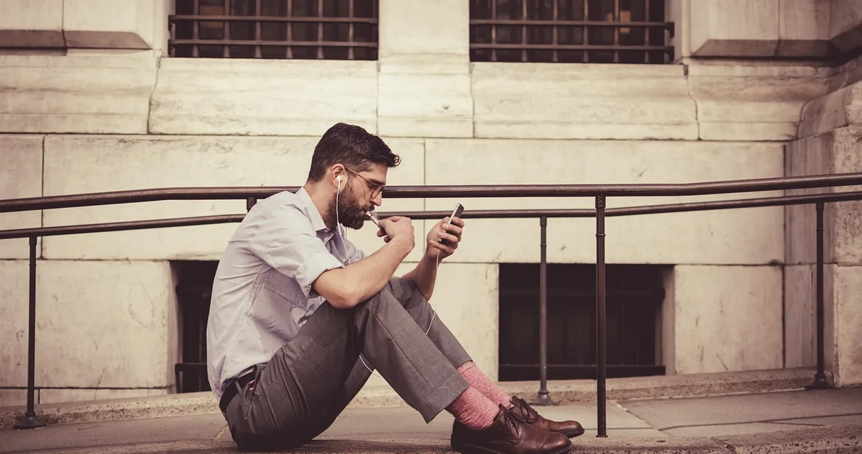Homem sentado na calçada usando o celular com fones de ouvido, ilustrando as notícias ao vivo no celular