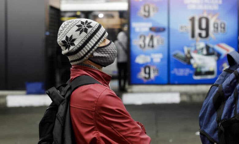 A imagem mostra uma pessoa de perfil, usando um gorro com padrões em preto e branco e uma máscara facial. A pessoa está vestindo uma jaqueta vermelha e parece estar em um ambiente urbano, com um fundo que apresenta um cartaz publicitário com preços e produtos. O ambiente é iluminado e parece ser uma área de comércio.