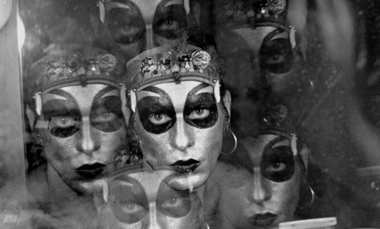 A imagem apresenta um retrato em preto e branco de uma pessoa com maquiagem dramática, incluindo olhos destacados em preto e lábios escuros. A figura parece refletida em uma superfície, criando um efeito de múltiplas camadas. O fundo é desfocado, e há um leve brilho que sugere um ambiente de backstage ou de performance.