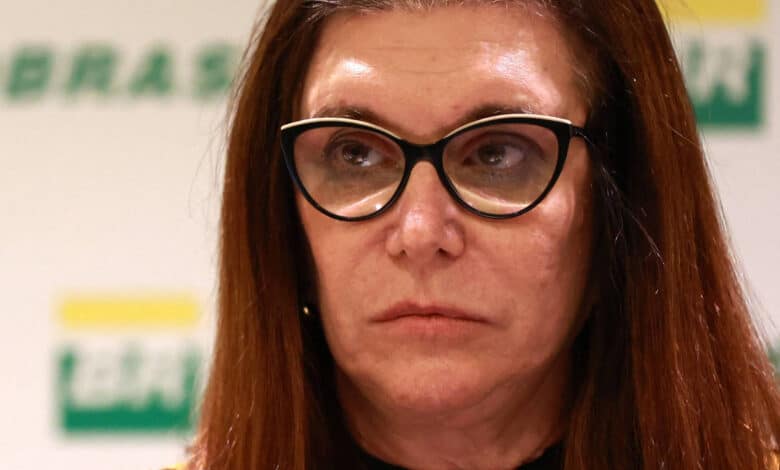 A imagem mostra uma mulher com cabelo longo e castanho, usando óculos grandes e escuros. Ela tem uma expressão séria e está olhando para frente. Ao fundo, há uma parede com a palavra