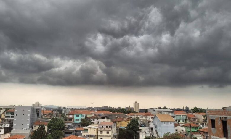 A imagem mostra uma vista panorâmica de uma cidade sob um céu nublado e escuro. As nuvens são densas e ameaçadoras, cobrindo a maior parte do céu. Abaixo, pode-se ver uma variedade de edifícios e casas com telhados de diferentes cores, predominantemente em tons de terracota e cinza. O horizonte é visível ao fundo, com algumas estruturas mais altas se destacando.