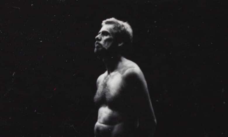 A imagem mostra um homem em pé, com o tronco nu e cabelo curto, olhando para cima. A iluminação é dramática, destacando seu rosto e parte do corpo, enquanto o fundo é escuro. A expressão do homem é séria e contemplativa, e a imagem tem um efeito de alto contraste, com predominância de tons escuros.