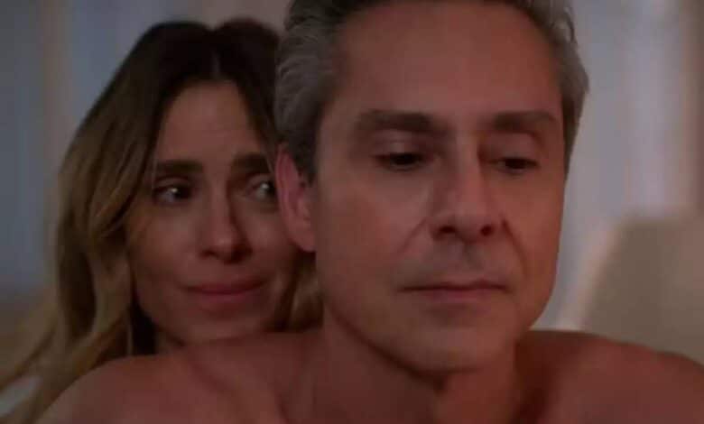 A imagem mostra um homem e uma mulher em um ambiente íntimo. O homem está sem camisa, com uma expressão séria, enquanto a mulher está atrás dele, com uma expressão de preocupação ou tristeza. O fundo é suave e desfocado, sugerindo um ambiente acolhedor.