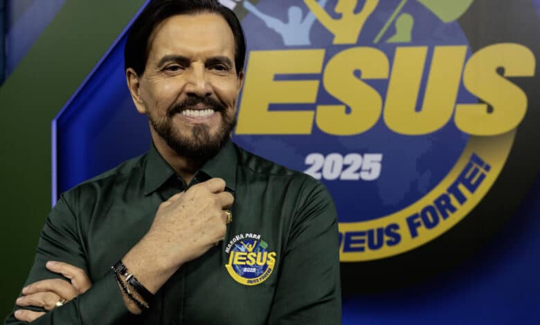 Um homem sorridente está posando para a foto em um estúdio. Ele usa uma camisa verde escura e está com a mão no queixo. Ao fundo, há um logotipo colorido que diz