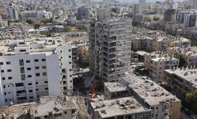 A imagem mostra uma área urbana devastada, com prédios danificados e em ruínas. Um edifício em destaque está parcialmente destruído, com escombros visíveis ao seu redor. Ao fundo, há uma vista da cidade com vários arranha-céus e construções. O céu está claro e a cena parece ser de um local que sofreu um desastre, possivelmente um ataque ou um terremoto.