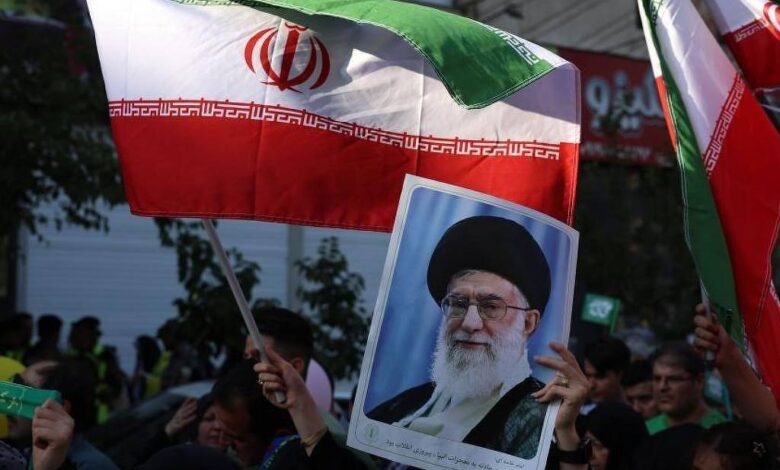 A imagem mostra um grupo de pessoas em um protesto, segurando bandeiras do Irã. Algumas pessoas também estão segurando um retrato de um líder iraniano. O ambiente parece ser urbano, com edifícios ao fundo e uma atmosfera de manifestação.