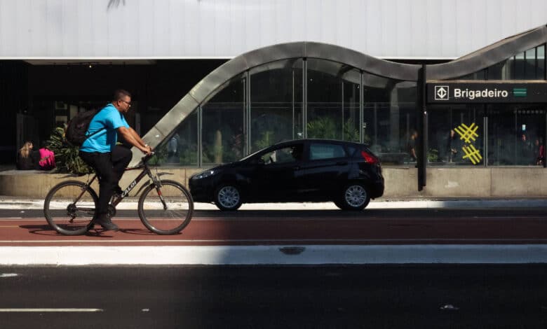 A imagem mostra um ciclista pedalando em uma ciclovia, ao lado de uma rua onde um carro preto está estacionado. Ao fundo, há uma estrutura arquitetônica com um design curvo e moderno, e uma estação de metrô com o nome