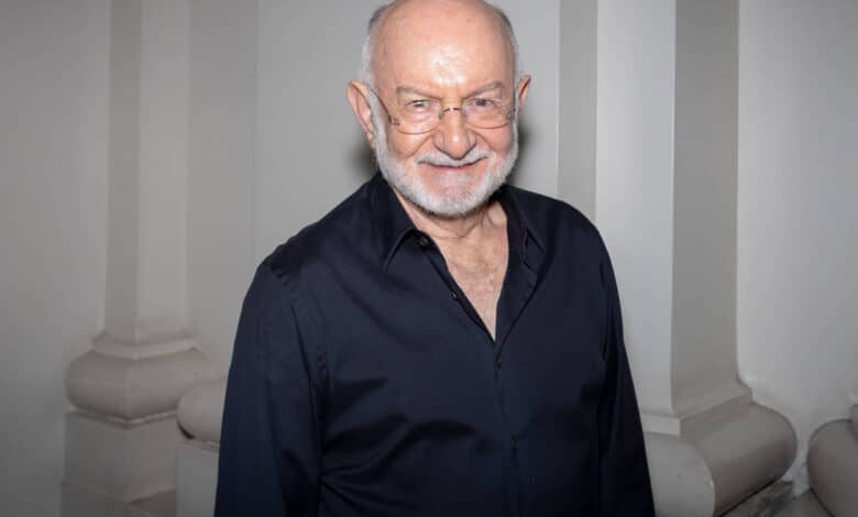 Um homem idoso com cabelo grisalho e barba branca, usando óculos e uma camisa preta, está sorrindo em um ambiente com paredes claras e colunas. Ele parece estar posando para a foto.