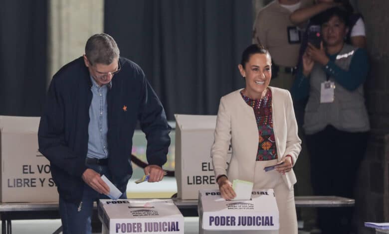 A imagem mostra duas pessoas participando de uma votação. À esquerda, um homem com cabelo curto e vestindo uma jaqueta azul escura e uma camiseta clara, está colocando um voto em uma urna. À direita, uma mulher com cabelo preso, vestindo um traje claro e uma blusa colorida, sorri enquanto segura um papel na mão. Ao fundo, outras pessoas observam a cena. As urnas têm a inscrição