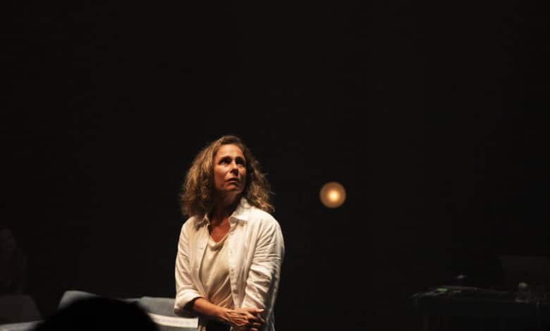 Uma mulher com cabelo cacheado e solto, vestindo uma camisa branca, está em um palco com iluminação suave. Ela parece estar em um momento de reflexão ou emoção, olhando para o lado. O fundo é escuro, com uma luz amarelada visível ao fundo, e há alguns objetos de cena, como cadeiras, em um ambiente teatral.