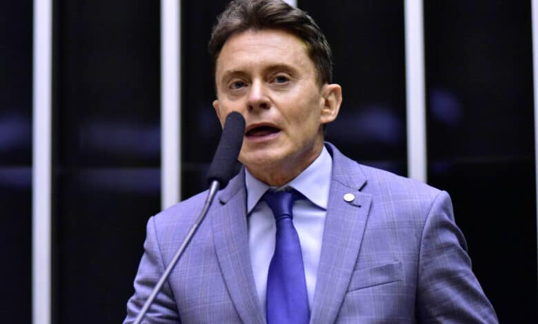 Um homem em um terno cinza claro e gravata azul está falando em um microfone em um ambiente de parlamento. Ele parece estar em uma posição de destaque, com um fundo que inclui painéis verticais. O homem tem cabelo castanho e está expressando-se de forma enérgica.