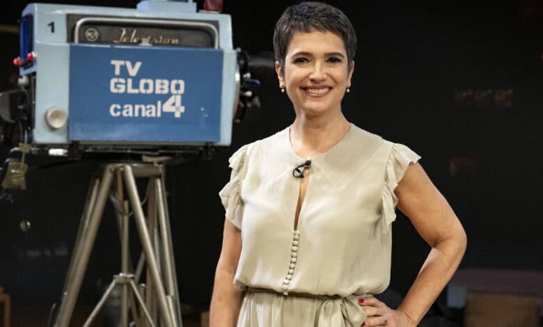 Uma mulher sorridente está em pé em um estúdio de televisão, vestindo um vestido claro. Ao fundo, há uma câmera de TV antiga com a inscrição