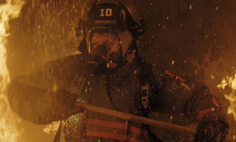 Um bombeiro está lutando contra um incêndio em um ambiente cheio de chamas e fumaça. Ele usa um capacete com o número 10 e uma máscara de proteção, segurando uma ferramenta de combate a incêndios. O fundo é dominado por fogo e fumaça, criando uma atmosfera intensa e perigosa.
