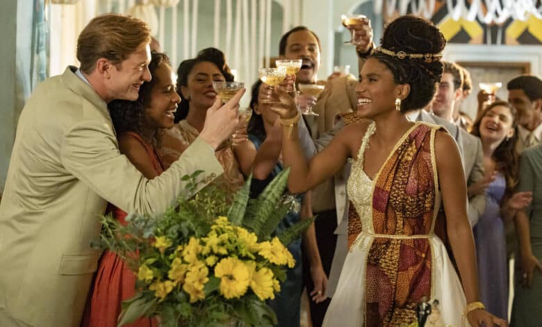 A imagem mostra um grupo de pessoas em uma festa, levantando taças em um brinde. No centro, uma mulher com um vestido branco e detalhes coloridos sorri enquanto brinda. Ao seu redor, há homens e mulheres de diferentes etnias, todos sorrindo e celebrando. O ambiente é decorado com flores amarelas e há uma atmosfera festiva.