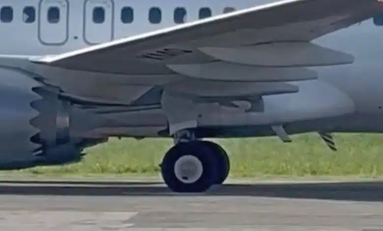 A imagem mostra a parte inferior de um avião, focando na roda do trem de pouso. O avião é de cor cinza e está posicionado em uma pista de pouso, com grama verde ao fundo. As asas do avião são visíveis, assim como parte do motor.
