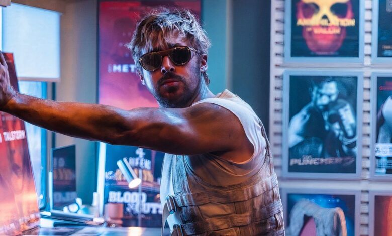 Ryan Gosling wearing sunglasses and a dirty vest stands in a room full of movie posters in The Fall Guy.