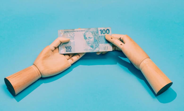 A imagem mostra duas mãos de madeira, uma de cada lado, segurando uma nota de 100 reais. O fundo é azul claro, destacando a nota que possui a figura de uma mulher e o valor impresso em destaque. As mãos estão posicionadas de forma a parecer que estão trocando a nota.