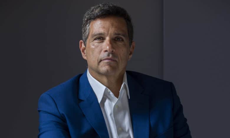 Um homem está sentado à mesa, vestindo um terno azul e uma camisa branca. Ele tem cabelo curto e grisalho, e está olhando para a câmera com uma expressão séria. A mesa reflete sua imagem, criando um efeito visual interessante. O fundo é de cor neutra, destacando o sujeito da imagem.