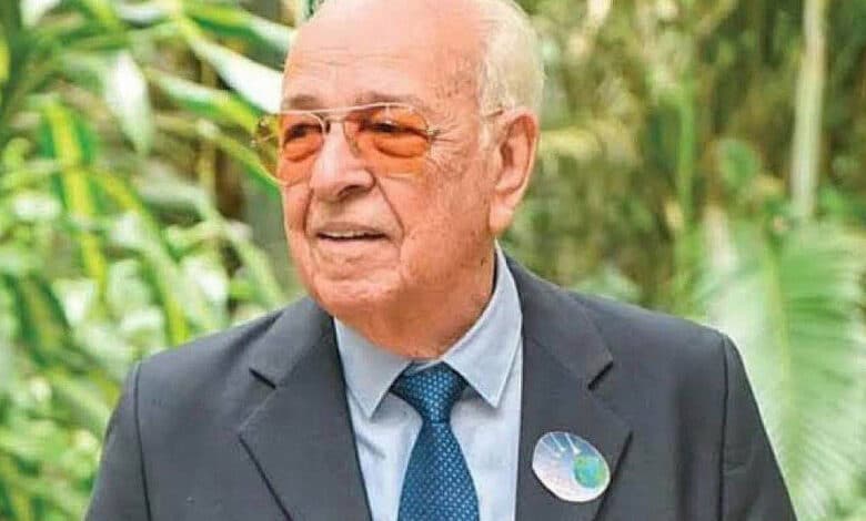 Um homem idoso está posando em um ambiente natural, cercado por plantas verdes. Ele usa um terno escuro, uma camisa clara e óculos de sol laranja. Na lapela do seu paletó, há um botão com um desenho que parece representar o planeta Terra.