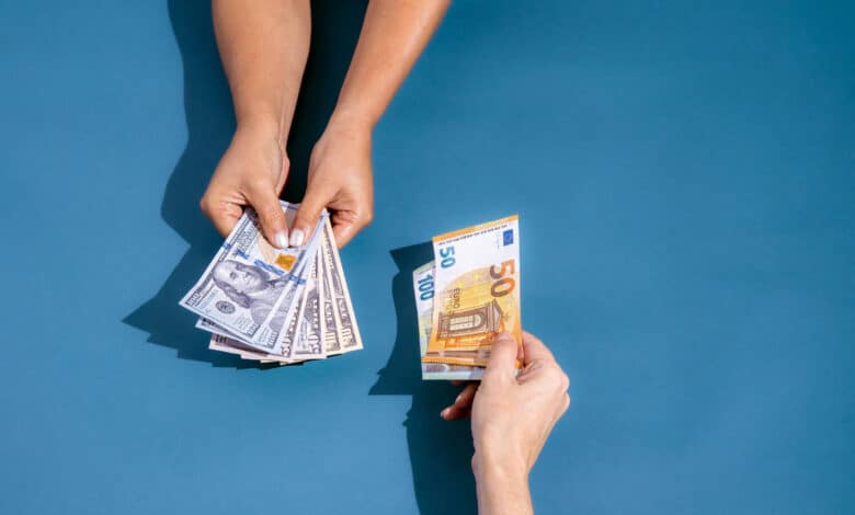 A imagem mostra duas mãos em um fundo azul. Uma mão está segurando várias notas de dólar, enquanto a outra mão está segurando uma nota de cinquenta euros. As mãos estão se aproximando, sugerindo uma troca de dinheiro.