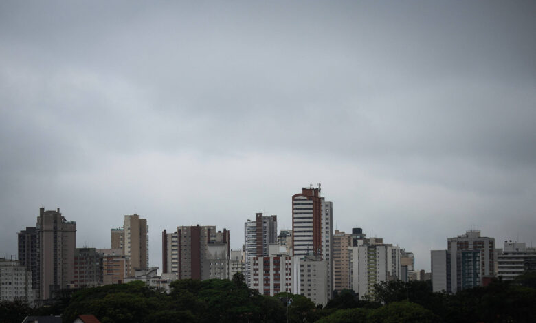 A imagem mostra uma paisagem urbana com vários edifícios altos em um dia nublado. O céu está coberto por nuvens cinzentas, criando uma atmosfera sombria. Os prédios são de diferentes tamanhos e estilos arquitetônicos, e há uma vegetação visível na parte inferior da imagem, sugerindo a presença de árvores ou parques na área.
