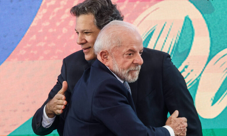 Na imagem, duas pessoas estão se abraçando em um evento. Um homem está à esquerda, com cabelo castanho e liso, vestindo um terno escuro. O homem à direita tem cabelo grisalho e uma barba curta, também vestindo um terno escuro. Ao fundo, há uma tela colorida com padrões vibrantes.