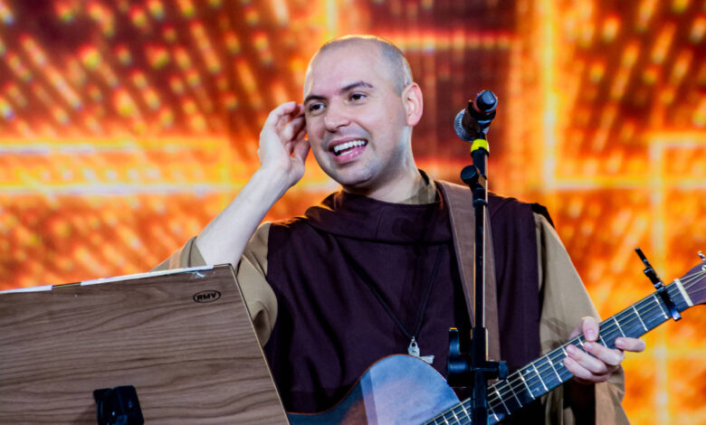 Um homem sorridente, vestido com um hábito marrom, está em um palco segurando um violão. Ele parece estar se apresentando, com um microfone próximo à boca e uma partitura em um suporte ao seu lado. O fundo é iluminado com luzes brilhantes em tons quentes, criando uma atmosfera vibrante.