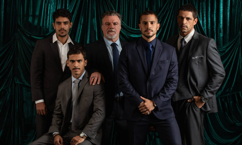 A imagem mostra cinco homens em trajes formais, posando em frente a um fundo de veludo verde. Os homens estão em diferentes poses: um está sentado, enquanto os outros estão em pé. Eles usam ternos de cores variadas, incluindo cinza e azul escuro, e todos têm expressões sérias. O ambiente é bem iluminado, destacando os detalhes dos trajes e o fundo texturizado.