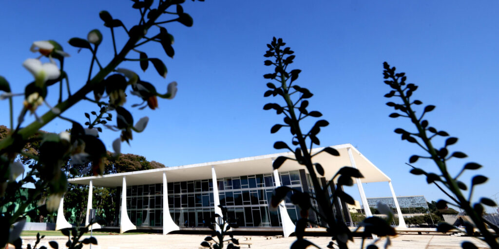 Em primeiro plano, silhueta de folhas em frente ao jardim do edifício sede do STF, que aparece ao fundo. Dia claro de céu azul.