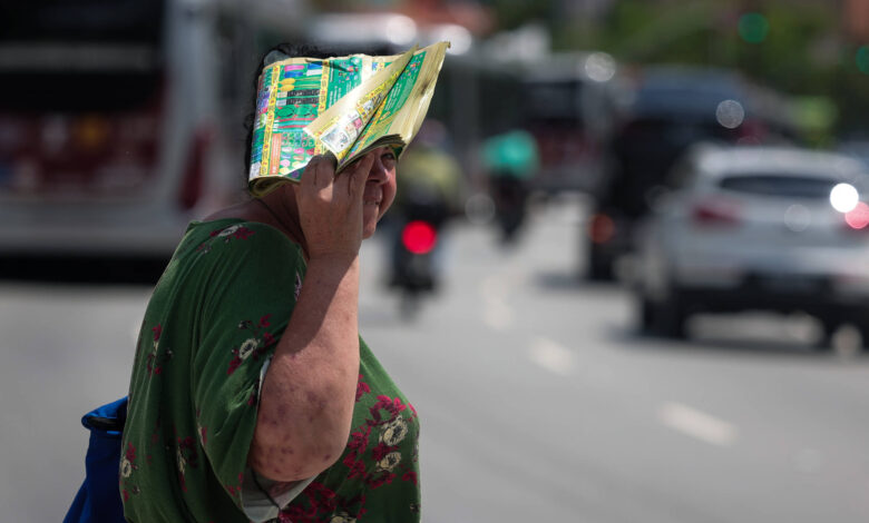 Uma mulher está na rua segurando um mapa sobre a cabeça, como se estivesse se protegendo do sol. Ela usa uma blusa verde com estampas florais e uma saia vermelha. Ao fundo, há veículos em movimento, incluindo ônibus e carros, e semáforos visíveis.