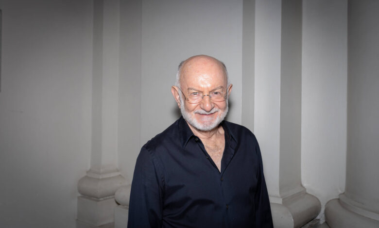 Um homem idoso com cabelo grisalho e barba branca, usando óculos e uma camisa preta, está sorrindo em um ambiente com paredes claras e colunas. Ele parece estar posando para a foto.