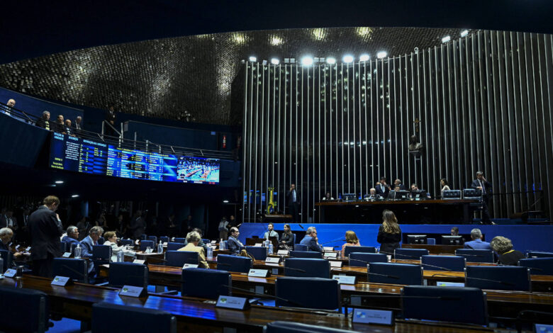 A imagem mostra o plenário do Senado Federal do Brasil, com senadores sentados em mesas dispostas em formato de U. Ao fundo, há uma tela de votação e um painel com informações. O ambiente é iluminado e possui um teto com design moderno. Algumas pessoas estão de pé, enquanto outras estão sentadas, e há uma bandeira do Brasil visível.