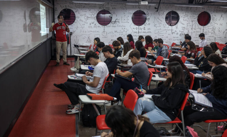 A imagem mostra uma sala de aula com um professor em pé à esquerda, apresentando um conteúdo em uma tela. Os alunos estão sentados em carteiras, a maioria com cadernos e canetas, prestando atenção ao professor. As paredes da sala estão cobertas com anotações e fórmulas matemáticas, e há janelas redondas ao fundo.