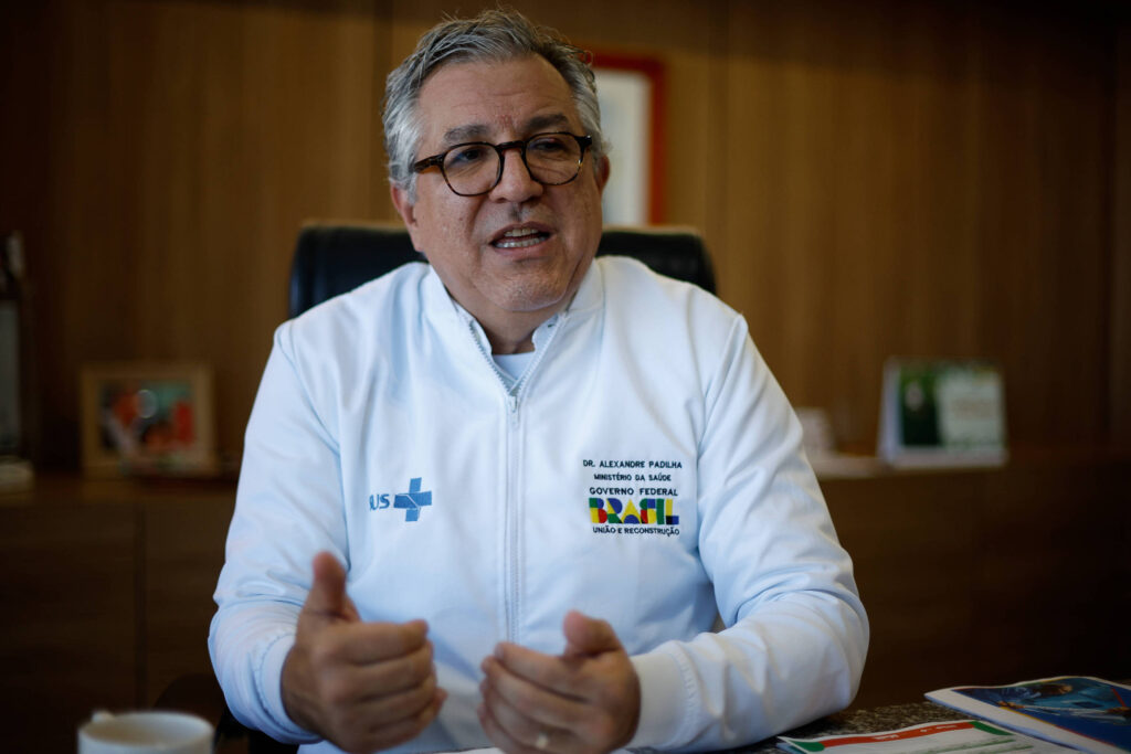 Imagem mostra o ministro Alexandre Padilha, um homem com cabelo grisalho e óculos está sentado em uma cadeira, usando um jaleco branco com o logotipo do SUS. Ele está gesticulando com as mãos enquanto fala. Ao fundo, há uma mesa de escritório com alguns objetos e documentos, além de uma parede de madeira.