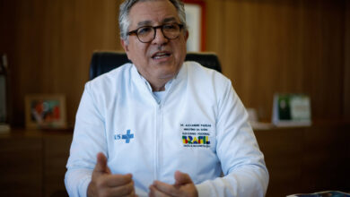 Imagem mostra o ministro Alexandre Padilha, um homem com cabelo grisalho e óculos está sentado em uma cadeira, usando um jaleco branco com o logotipo do SUS. Ele está gesticulando com as mãos enquanto fala. Ao fundo, há uma mesa de escritório com alguns objetos e documentos, além de uma parede de madeira.