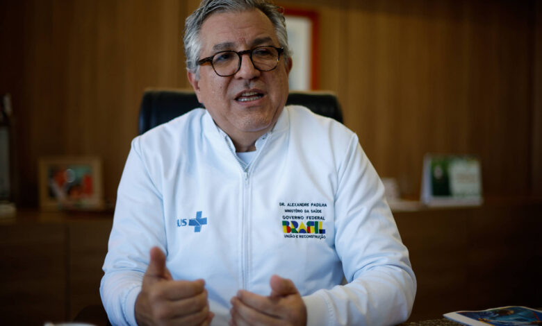 Imagem mostra o ministro Alexandre Padilha, um homem com cabelo grisalho e óculos está sentado em uma cadeira, usando um jaleco branco com o logotipo do SUS. Ele está gesticulando com as mãos enquanto fala. Ao fundo, há uma mesa de escritório com alguns objetos e documentos, além de uma parede de madeira.