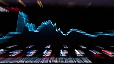 A imagem mostra um gráfico de linha em um fundo escuro, representando dados financeiros. O gráfico tem uma linha azul que sobe e desce, indicando flutuações nos valores. Abaixo do gráfico, há uma série de luzes coloridas que representam dados adicionais, possivelmente de um painel de controle ou terminal financeiro.