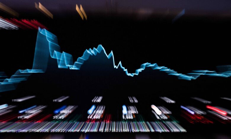 A imagem mostra um gráfico de linha em um fundo escuro, representando dados financeiros. O gráfico tem uma linha azul que sobe e desce, indicando flutuações nos valores. Abaixo do gráfico, há uma série de luzes coloridas que representam dados adicionais, possivelmente de um painel de controle ou terminal financeiro.