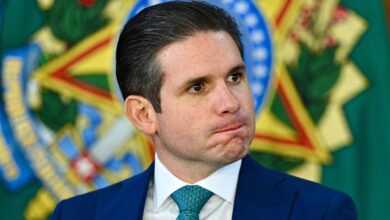 Um homem com cabelo liso e escuro, usando um terno azul e uma gravata verde, está sentado em um evento oficial. Ele tem uma expressão pensativa, com os lábios comprimidos. Ao fundo, há um emblema verde e dourado, que parece ser um símbolo nacional ou institucional.