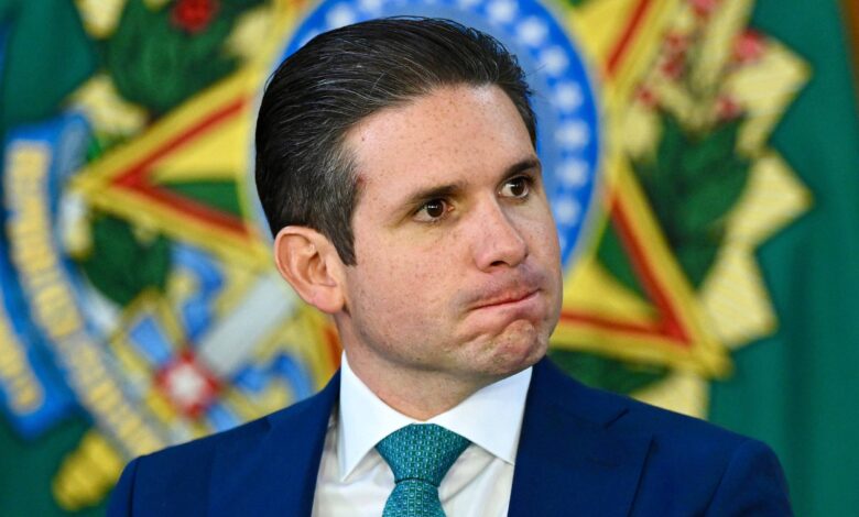Um homem com cabelo liso e escuro, usando um terno azul e uma gravata verde, está sentado em um evento oficial. Ele tem uma expressão pensativa, com os lábios comprimidos. Ao fundo, há um emblema verde e dourado, que parece ser um símbolo nacional ou institucional.
