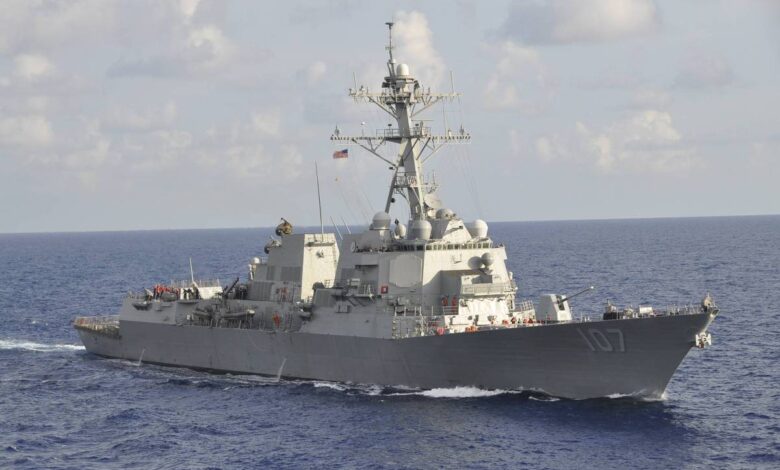 Um navio de guerra, possivelmente um destróier, navega em águas abertas. O navio é de cor cinza e possui vários equipamentos e antenas visíveis em sua estrutura. Há pessoas a bordo, e a bandeira dos Estados Unidos está hasteada no mastro. O céu está parcialmente nublado e o mar é calmo.