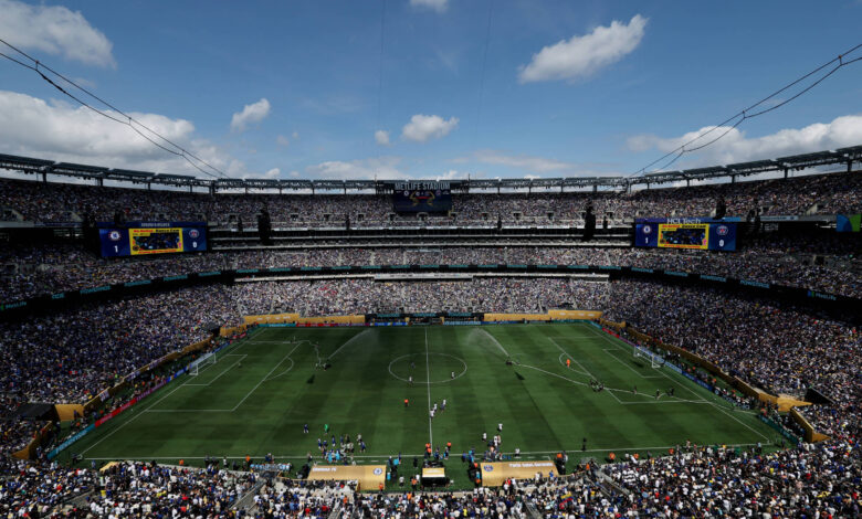 A imagem mostra um estádio de futebol lotado, com torcedores nas arquibancadas. O campo está visível, com jogadores em ação. O céu está parcialmente nublado, e há grandes telões visíveis nas laterais do estádio. A atmosfera é de grande expectativa e emoção, típica de um evento esportivo.
