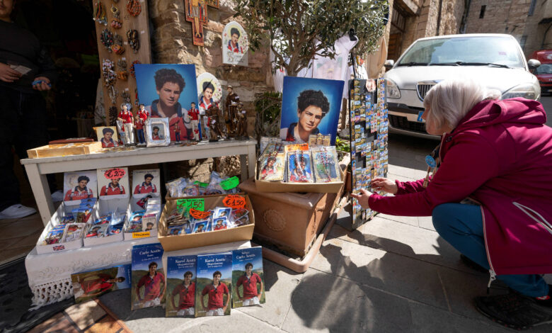 A imagem mostra uma cena de um mercado de artesanato ao ar livre. À esquerda, há uma mesa com diversos itens, incluindo ícones religiosos, fotos de pessoas e pequenos objetos decorativos. No chão, há pacotes com imagens e produtos variados. À direita, uma mulher com cabelo claro e uma jaqueta rosa está agachada, observando os produtos expostos. Ao fundo, é possível ver uma parede de pedra e um carro estacionado.