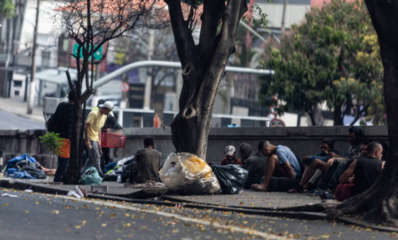 A imagem mostra um grupo de pessoas em situação de rua em um ambiente urbano. Algumas pessoas estão sentadas no chão, enquanto outras estão de pé. Há sacos de lixo e cobertores espalhados pelo chão. Árvores estão presentes ao redor, e ao fundo, é possível ver edifícios e uma rua. A iluminação sugere que é um dia claro.