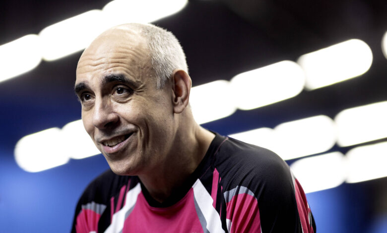 Um homem com cabelo grisalho e calvo, sorrindo, está usando uma camiseta esportiva de cor preta com detalhes em rosa e branco. O fundo é desfocado, com luzes brilhantes que criam um efeito de iluminação. O homem parece estar em um ambiente de competição, possivelmente relacionado a esportes.