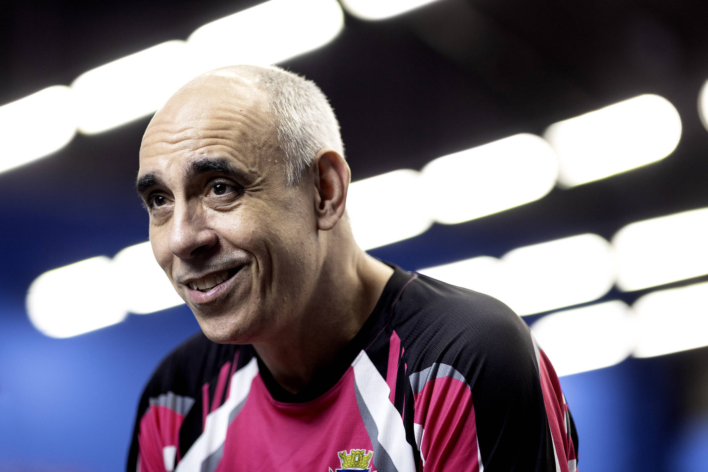 Um homem com cabelo grisalho e calvo, sorrindo, está usando uma camiseta esportiva de cor preta com detalhes em rosa e branco. O fundo é desfocado, com luzes brilhantes que criam um efeito de iluminação. O homem parece estar em um ambiente de competição, possivelmente relacionado a esportes.