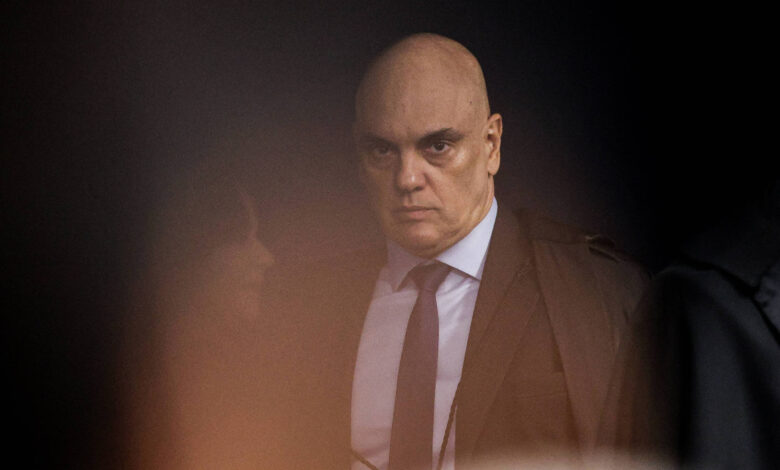 A imagem mostra um homem careca com uma expressão séria. Ele está vestido com um terno escuro e uma gravata. O fundo é desfocado, dificultando a visualização de outros detalhes. A iluminação é suave, criando um efeito de sombra ao redor do homem.