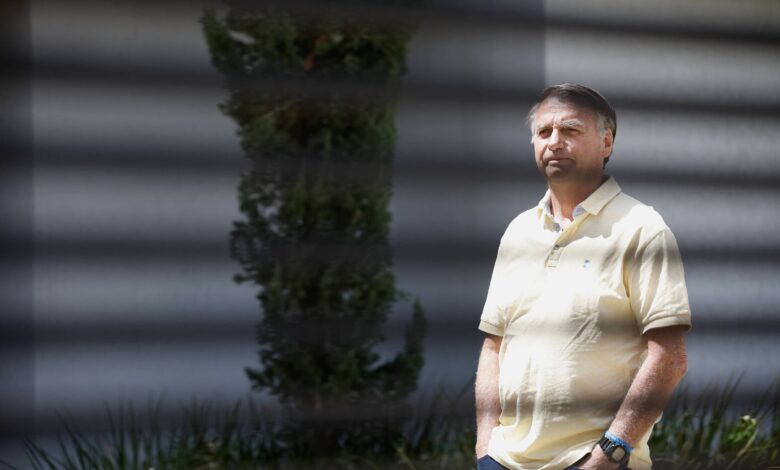 Um homem está em pé, olhando para frente, em um ambiente externo. Ele usa uma camisa polo amarela e calças escuras. Ao fundo, há uma planta verde e uma parede clara. Sombras de uma grade são visíveis na imagem, criando um efeito de listras sobre o homem.