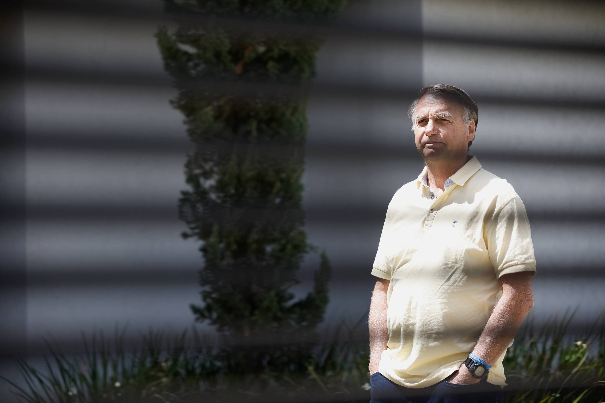 Um homem está em pé, olhando para frente, em um ambiente externo. Ele usa uma camisa polo amarela e calças escuras. Ao fundo, há uma planta verde e uma parede clara. Sombras de uma grade são visíveis na imagem, criando um efeito de listras sobre o homem.