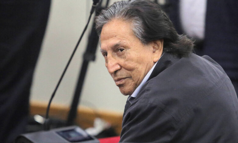 Um homem de cabelo longo e escuro, com um olhar pensativo, está sentado em um ambiente que parece ser uma audiência ou tribunal. Ele usa uma jaqueta escura e está voltado para o lado, com alguns microfones e câmeras visíveis ao fundo.